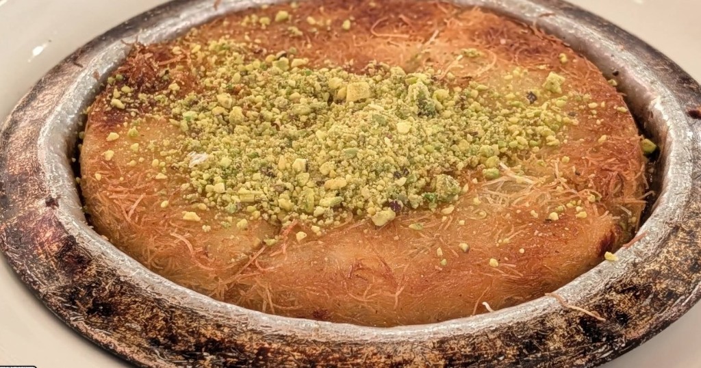 Golden kunefe dessert topped with crushed pistachios, served warm in a copper pan